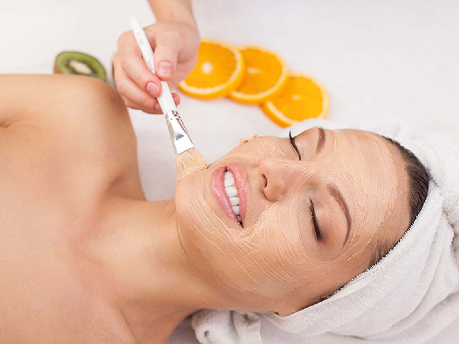 A person relaxing with a towel on their head enjoys a Vitamin C facial mask treatment, with slices of fruit nearby adding a refreshing touch.