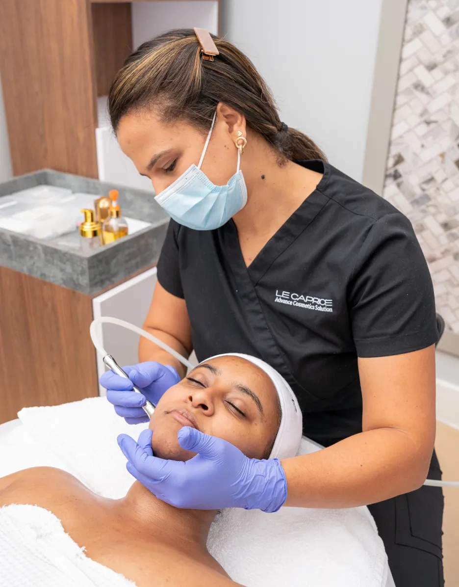 Esthetician in scrubs and mask performing a facial treatment on a client lying on a treatment bed.